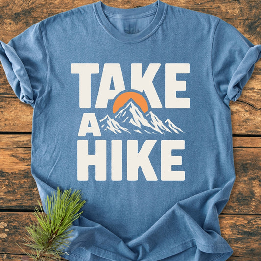 Take A Hike - Vintage Wash Tee featuring a heather-blue color with a bold “TAKE A HIKE” print, stylized mountain range, and orange sun. Laid on wooden surface with a pine branch beside it.