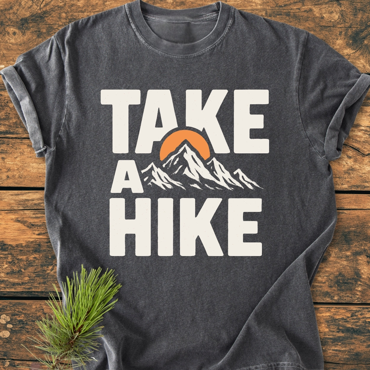 Take A Hike - Vintage Wash Tee features a stylized mountain graphic with “TAKE A HIKE” text, laid on a wooden surface with a pine sprig, showcasing its relaxed fit and durable stitching.