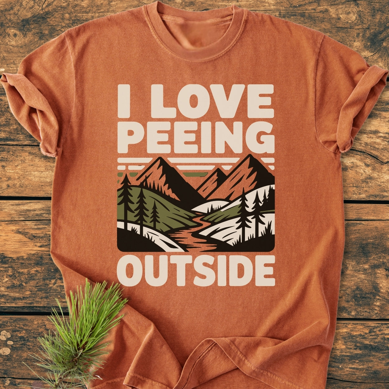 I Love Peeing Outside - Vintage Wash Tee featuring a stylized outdoor scene with mountains and trees, laid flat on a wooden surface with a pine sprig beside it.