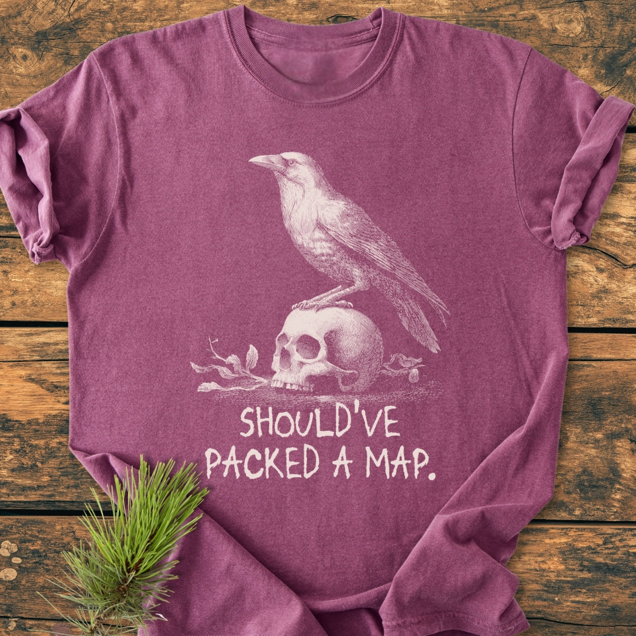 No WI-FI - Vintage Wash Tee: A folded T-shirt featuring a bird standing on a skull with leaves, paired with the slogan “SHOULD’VE PACKED A MAP” on a wooden surface.