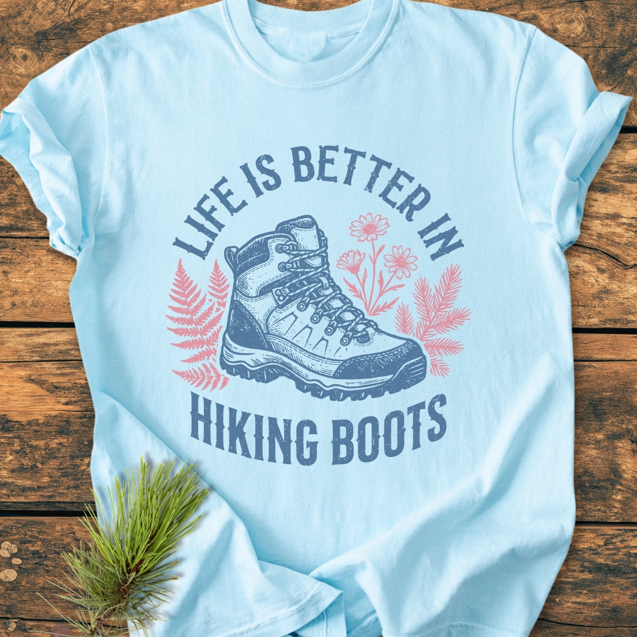 Life Is Better - Vintage Wash Tee featuring a hiking boot graphic with fern and flower illustrations, laid on wood, showcasing the phrase “LIFE IS BETTER IN HIKING BOOTS.” Pine sprig accentuates natural theme.