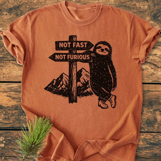 Not Fast Not Furious - Vintage Wash Tee featuring a cartoon sloth with hiking boots and a signpost. Laid on wood with pine sprig, showcasing the relaxed fit and durable stitching.