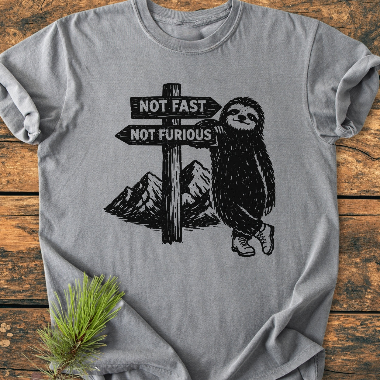Not Fast Not Furious - Vintage Wash Tee: A heather-gray T-shirt featuring a black graphic of a smiling sloth in boots, leaning on a signpost with playful arrows, laid on a wooden surface.