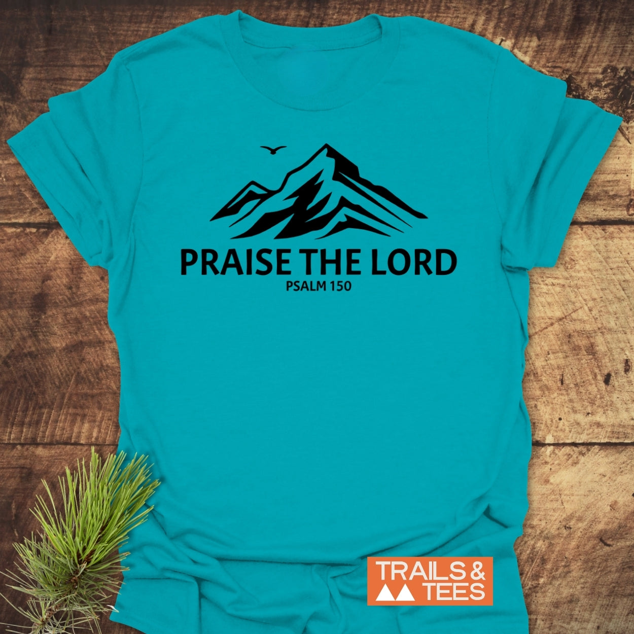 Praise The Lord, Psalm 150 T-Shirt featuring a mountain silhouette with a bird above on teal fabric, laid on wood with pine sprig; includes Trails & Tees logo in corner.