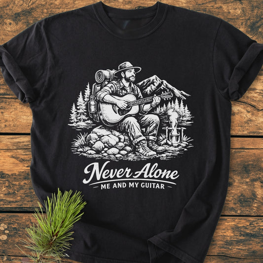 Never Alone. Me And My Guitar - Vintage Wash Tee features a bearded man by a campfire, playing guitar amid mountains and pines, printed on a soft, relaxed-fit, black T-shirt.