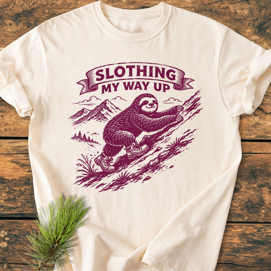 Slothing My Way Up - Vintage Wash Tee features a stylized sloth in hiking boots on a cream-colored T-shirt, laid out on wood, showcasing its durable, relaxed fit design.