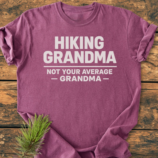 Heather-purple Hiking Grandma - Vintage Wash Tee with bold white text, laid flat on wood. Features soft-washed, garment-dyed cotton for comfort and durability, with a small pine sprig accent.