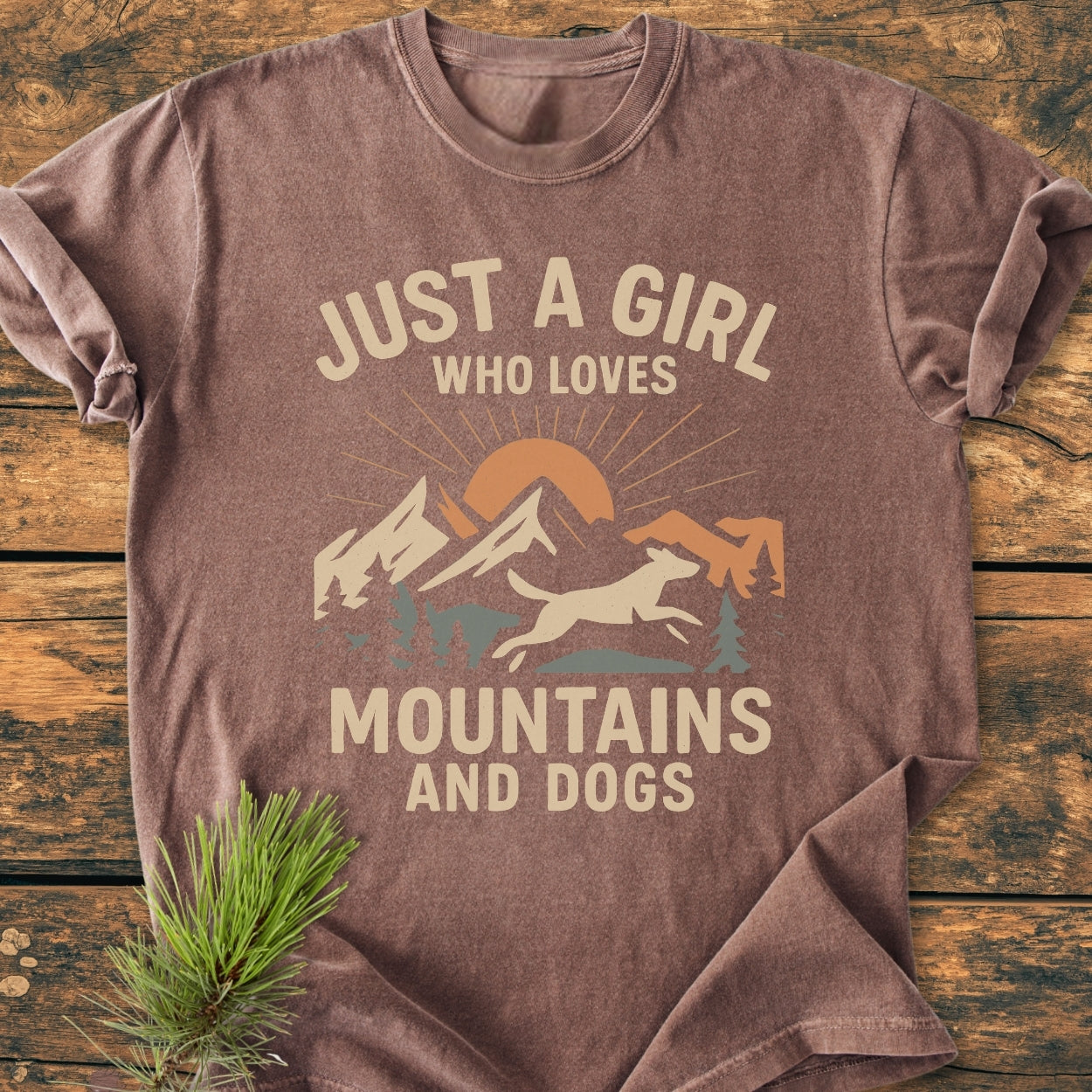Just A Girl Who Loves Mountains And Dogs - Vintage Wash Tee: Soft brown T-shirt with a mountain and leaping dog graphic, cream-and-muted-orange text on front, displayed on rustic wood with a pine sprig accent.