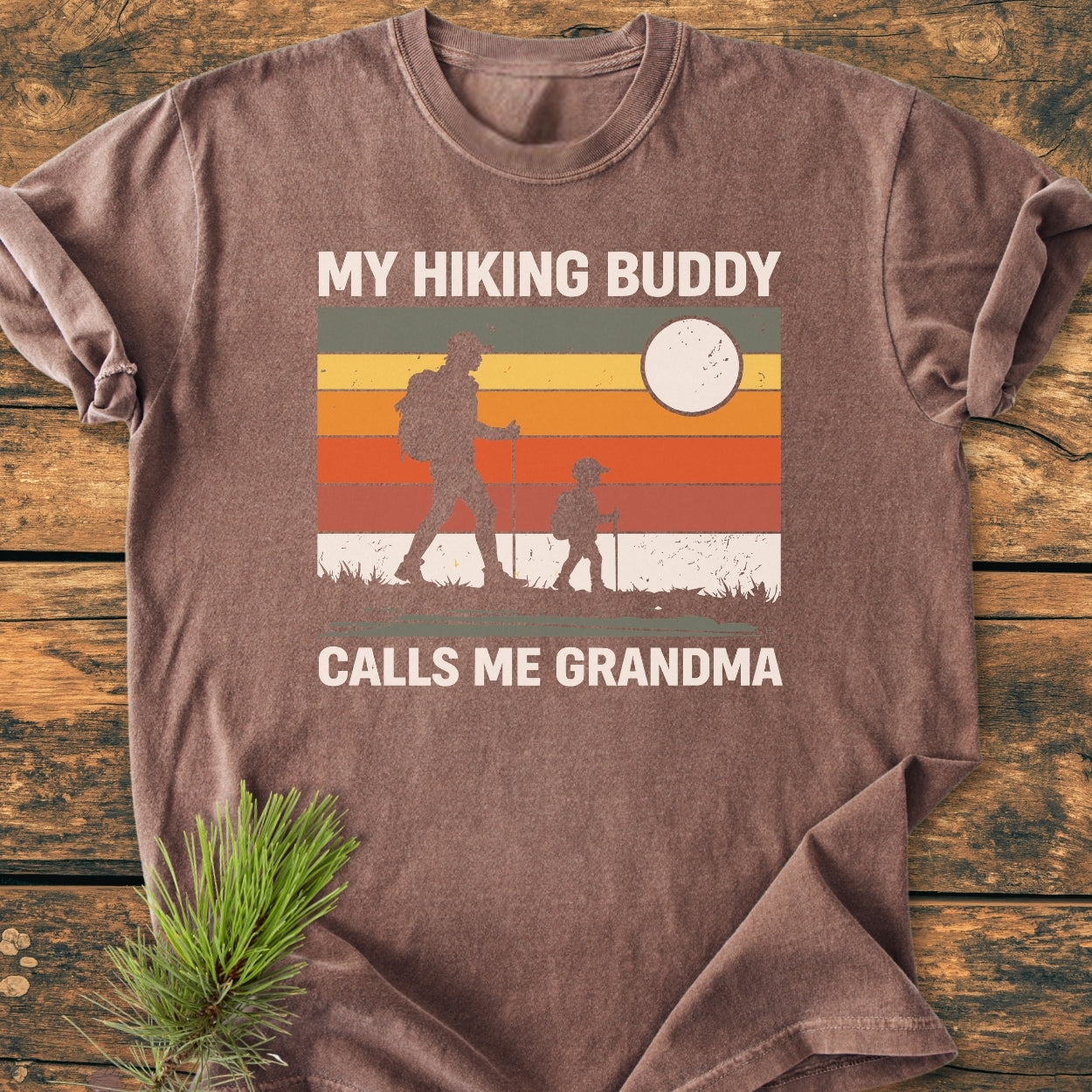 Vintage-style tee titled My Hiking Buddy Calls Me Grandma, featuring hikers against sunset stripes. Crafted from soft, garment-dyed cotton, offering a relaxed fit and durable stitching.