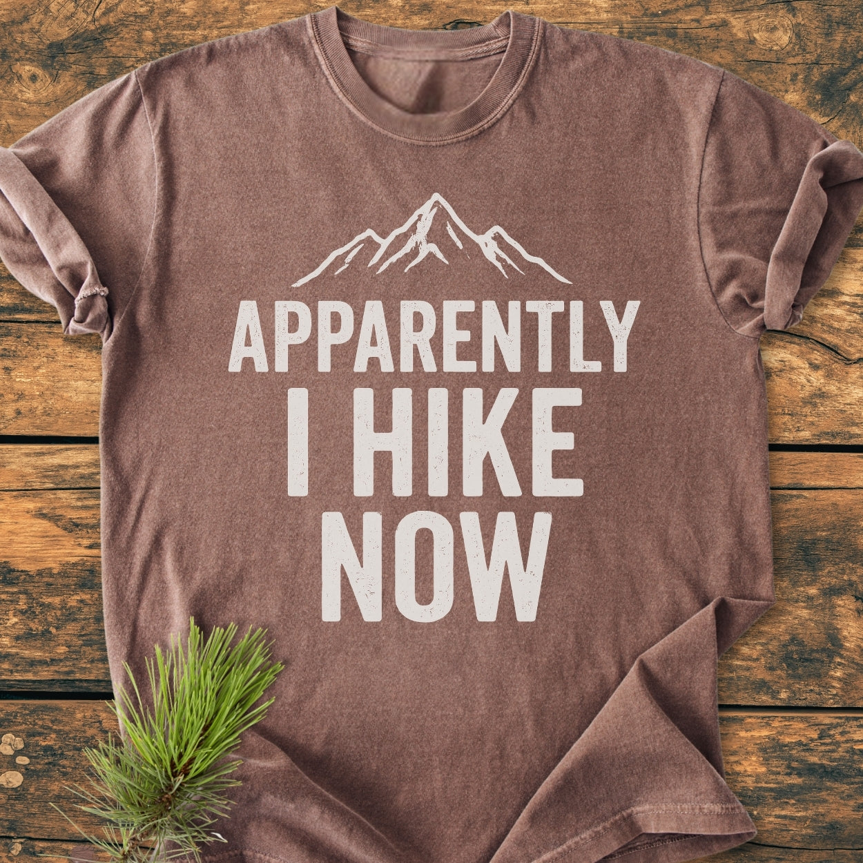 Heathered brown Apparently Hiking - Vintage Wash Tee with mountain peak graphic and phrase APPARENTLY I HIKE NOW in white, laid on wood surface with pine sprig accenting the scene.