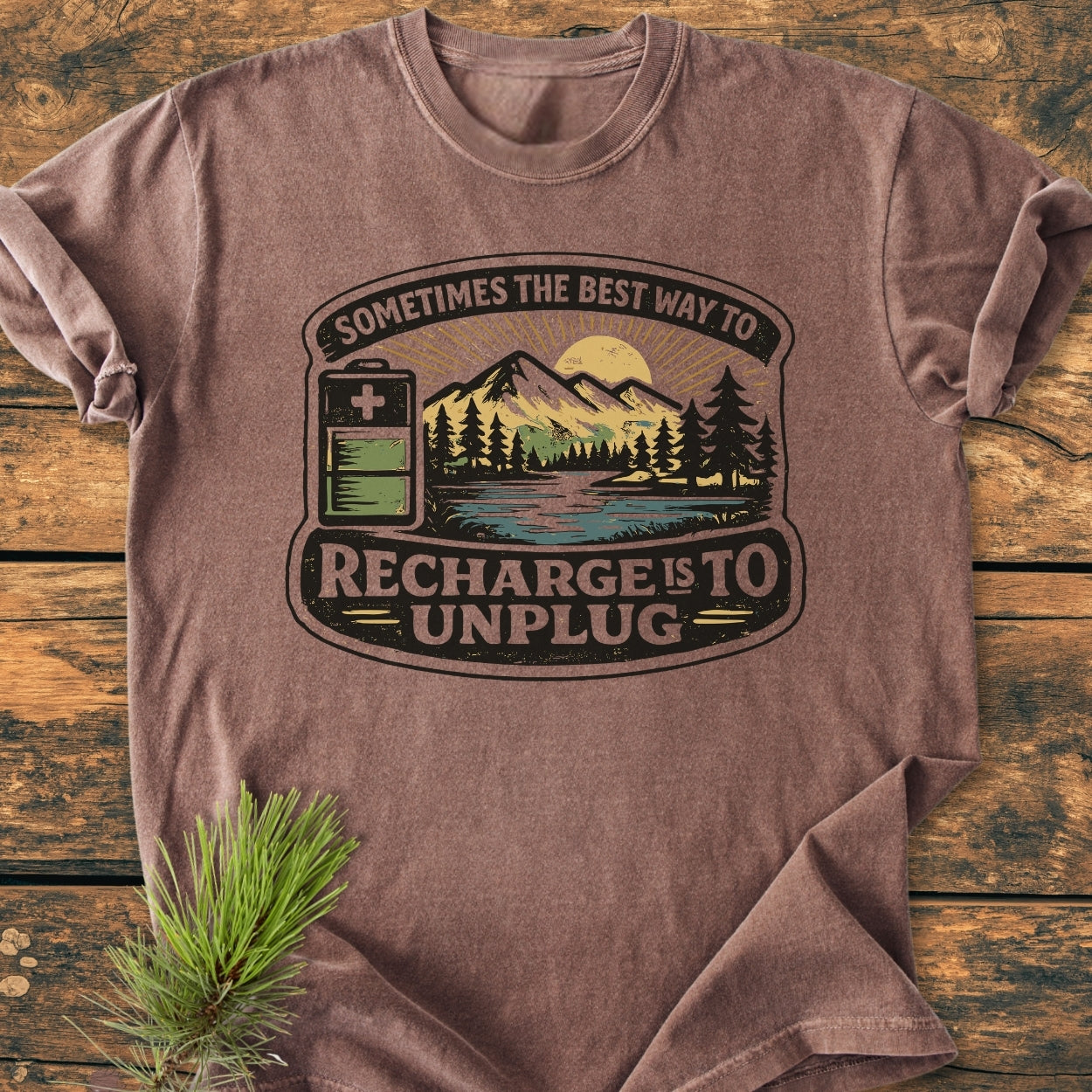 Unplug To Recharge - Vintage Wash Tee featuring a stylized battery and mountain scene with slogan, laid on wood with pine sprig, highlighting its soft, durable, relaxed-fit design in garment-dyed cotton.