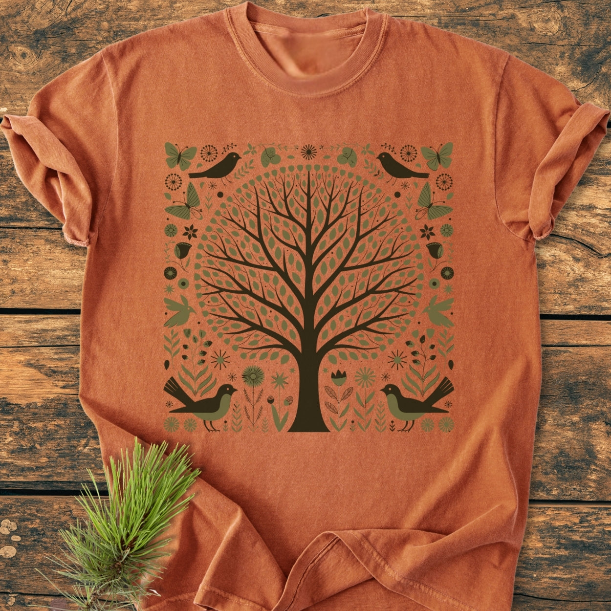 Nature Love - Vintage Wash Tee features a tree of life graphic with folk-art motifs. The orange-rust T-shirt is styled on a wooden surface, highlighting its relaxed fit and garment-dyed fabric.