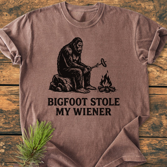Bigfoot Stole My Wiener - Vintage Wash Tee features a black-ink illustration of Bigfoot by a campfire on a brown T-shirt, capturing a playful outdoor scene. Made from soft, durable ring-spun cotton.