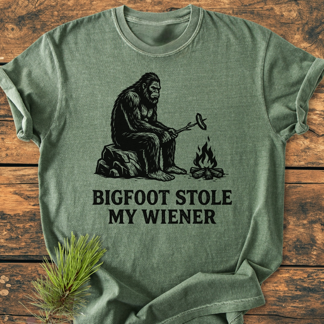 Bigfoot Stole My Wiener - Vintage Wash Tee features Bigfoot with a campfire graphic and humorous caption on a green Comfort Colors T-shirt, styled with a pine sprig on a wooden surface.