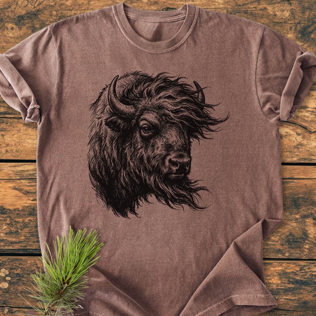 Silent Thunder - Vintage Wash Tee displays a black line-art bison head on a brown short-sleeve shirt. Laid on wood, a pine sprig accents its soft, relaxed fit, emphasizing comfort and durability.