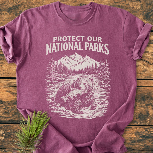 Protect Our National Parks - Vintage Wash Tee features a heather-purple design with a bear illustration and slogan, laid on wood. Soft, garment-dyed cotton ensures comfort and durability.