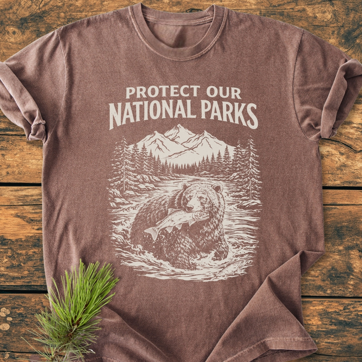 Protect Our National Parks - Vintage Wash Tee features a bear illustration with a fish, surrounded by nature. This brown, 100% cotton, relaxed fit t-shirt offers durability and comfort with double-needle stitching.