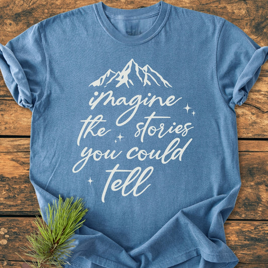 Imagine The Stories - Vintage Wash Tee features a heather-blue color with a white mountain graphic and slogan. Laid flat on wood, it includes a sprig of pine, highlighting its rustic charm.