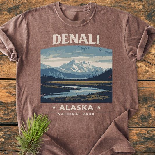 Denali National Park - Vintage Wash Tee features a mountain landscape with DENALI and ALASKA NATIONAL PARK text, laid on wood with a pine sprig, showcasing its relaxed fit and garment-dyed fabric.