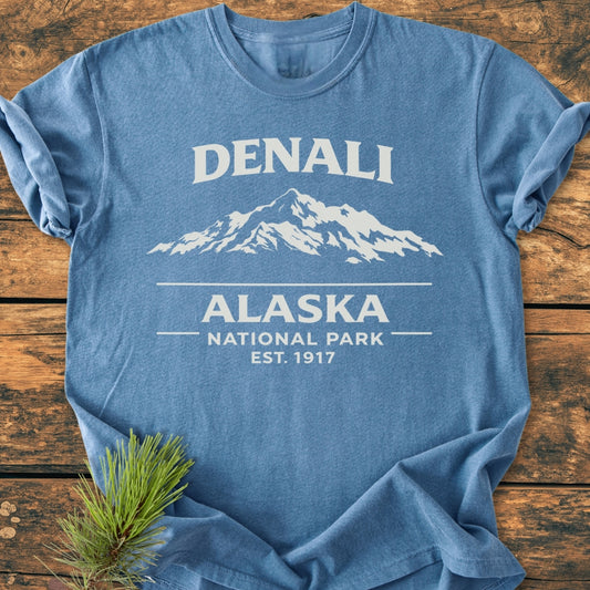 Denali Alaska National Park - Vintage Wash Tee featuring a mountain silhouette and text, laid on wood with a pine branch. Heather-blue, garment-dyed, 100% cotton, relaxed fit for outdoor appeal.