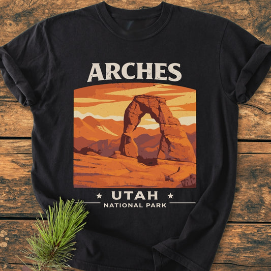 Arches Utah National Park - Vintage Wash Tee, featuring a retro graphic of Delicate Arch with ARCHES above. Laid on wood with a pine sprig, highlighting its cozy, relaxed fit and durable stitching.