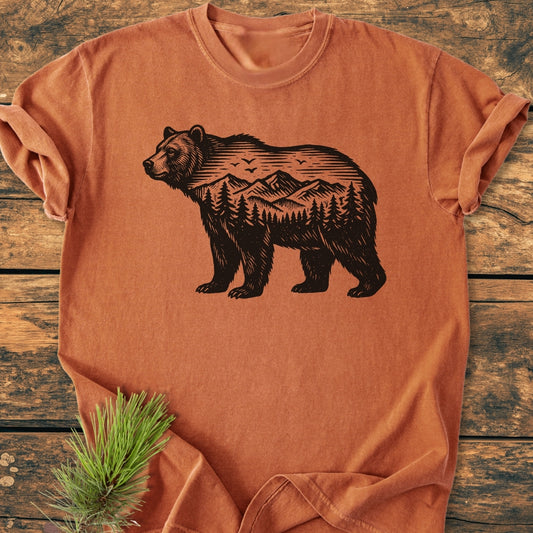 The Wilderness Within - Vintage Wash Tee features a black bear graphic with mountains and trees on a washed-rust-orange T-shirt, laid flat on a wooden surface with a pine sprig beside it.