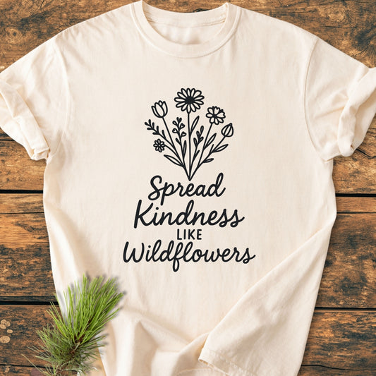 Spread Kindness - Vintage Wash Tee features a central line-drawing of wildflowers with the phrase “Spread Kindness Like Wildflowers” on an off-white tee, laid flat on a wooden surface.