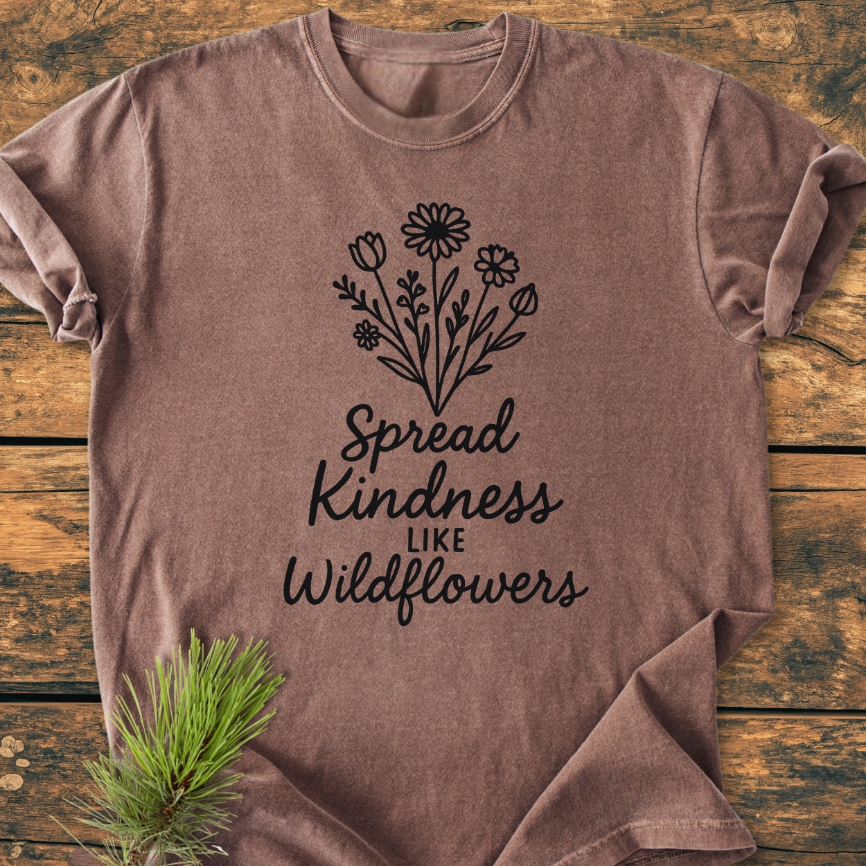 Spread Kindness - Vintage Wash Tee with black wildflower line-drawing and script text on heather-brown fabric, laid on wooden surface with pine branch styling prop, showcasing its relaxed fit and soft texture.