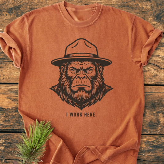 I Work Here - Vintage Wash Tee features a stylized Bigfoot face in a park-ranger hat with I WORK HERE text, laid on wood, a pine branch tucked under a corner.