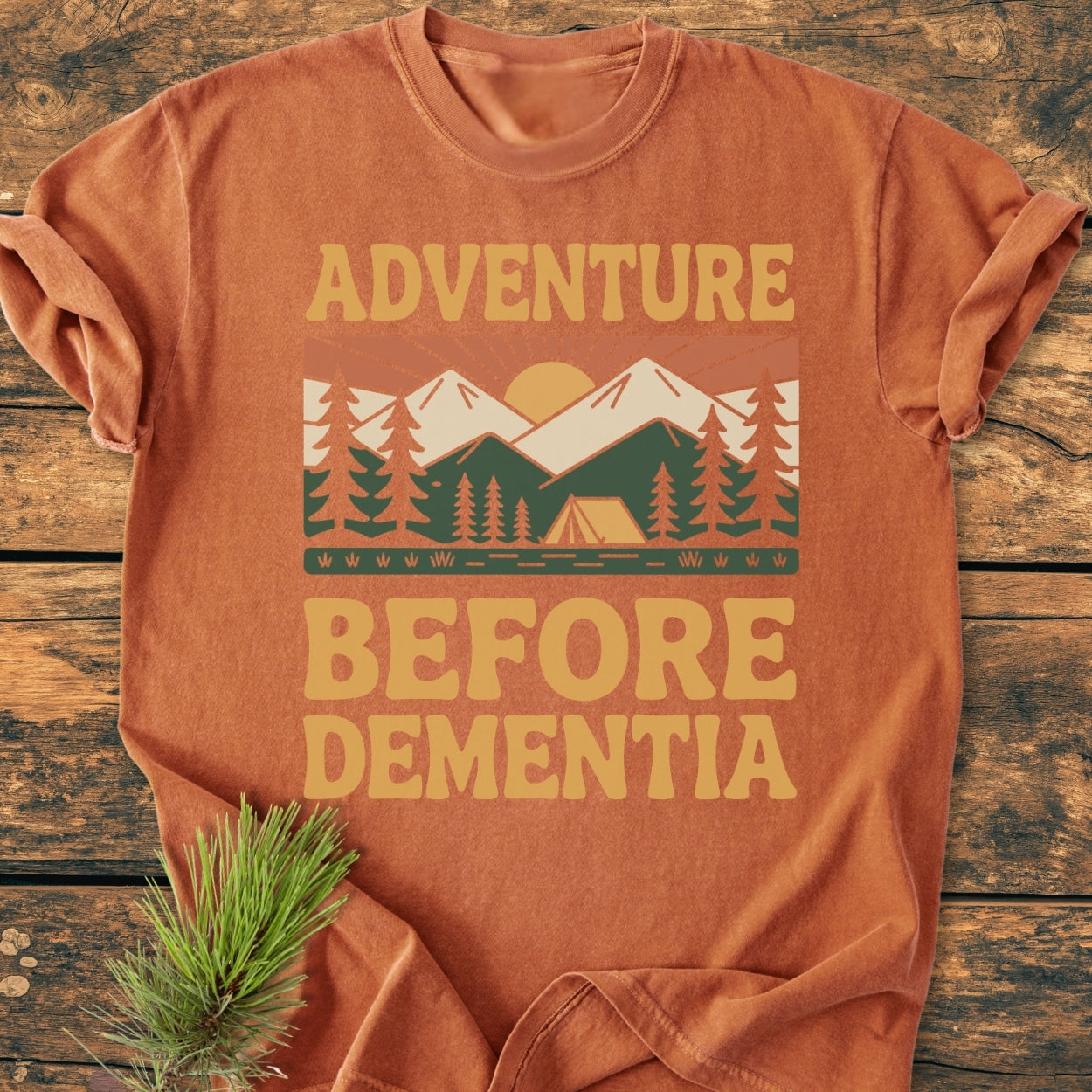 Adventure Before Dementia - Vintage Wash Tee featuring a stylized outdoor scene with pine trees, a tent, and mountains printed on a rust-orange T-shirt, laid on a wooden surface with a pine sprig.