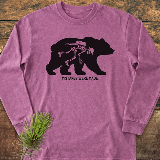 Mistakes Were Made Long-Sleeve Tee featuring a black bear silhouette with a hiker skeleton graphic, captioned below. Laid on wood, styled with a pine sprig, highlighting its relaxed, garment-dyed fit.