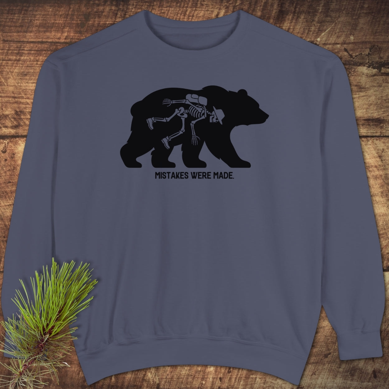 Mistakes Were Made Sweatshirt features a bear silhouette with human skeletal remains inside, captioned beneath. Displayed on rustic wood, emphasizing its luxurious, relaxed fit and garment-dyed fabric.