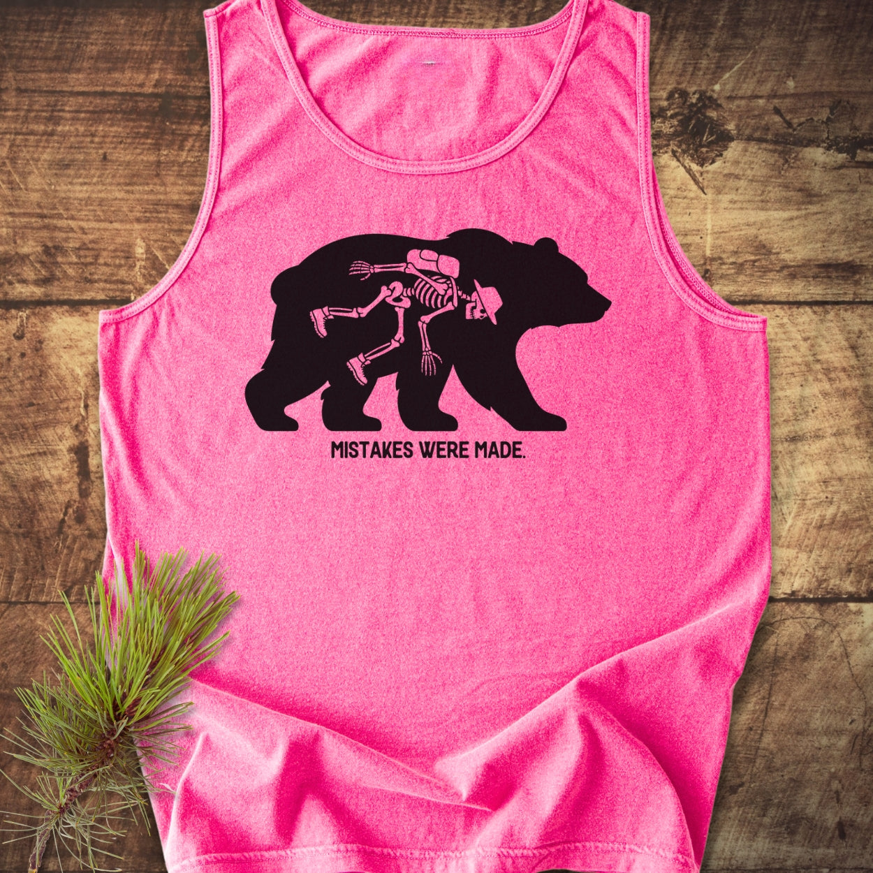 Mistakes Were Made Tank Top featuring a bear silhouette with a hiker skeleton print on a pink, sleeveless design, laid on wood. Offers relaxed fit, crafted from sustainable, ring-spun US cotton.