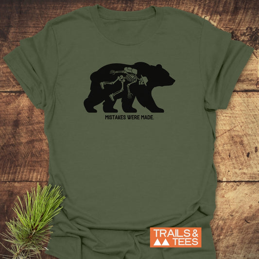 Mistakes Were Made T-Shirt featuring a bear silhouette with a hiker's bones inside, printed on an olive-green tee. Includes durable DTF prints, pre-shrunk fit, and styled with a pine branch.