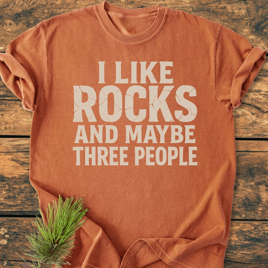 I Like Rocks - Vintage Wash Tee in rust-orange, featuring a cracked white slogan, styled with a pine sprig on weathered wood. Made from soft, pre-shrunk, ring-spun cotton with a relaxed fit.