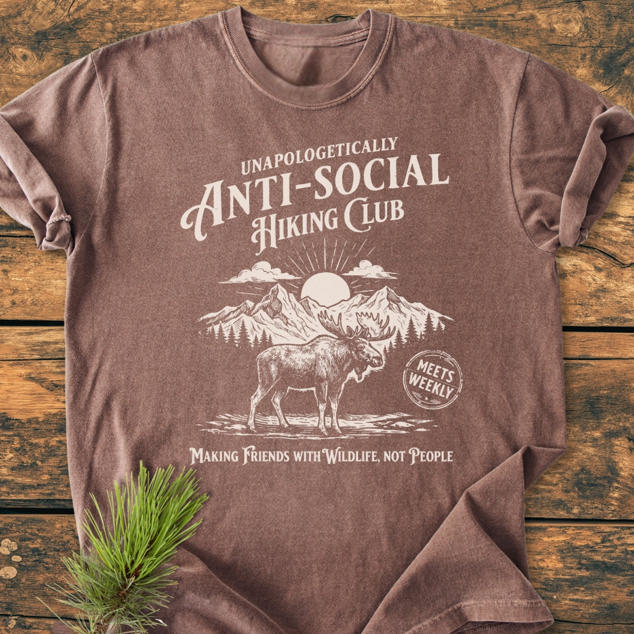 Anti-Social Hiking Club - Vintage Wash Tee folded on wood, featuring a moose and mountain illustration, Unapologetically Anti-Social, and Making Friends with Wildlife, Not People text. Pine sprig beside it.