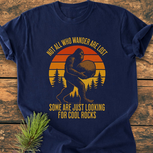 Looking For Cool Rocks - Vintage Wash Tee features Bigfoot carrying a stone with pine trees and a sunset backdrop. This comfortable, navy-blue tee offers a relaxed fit and durable double-needle stitching.
