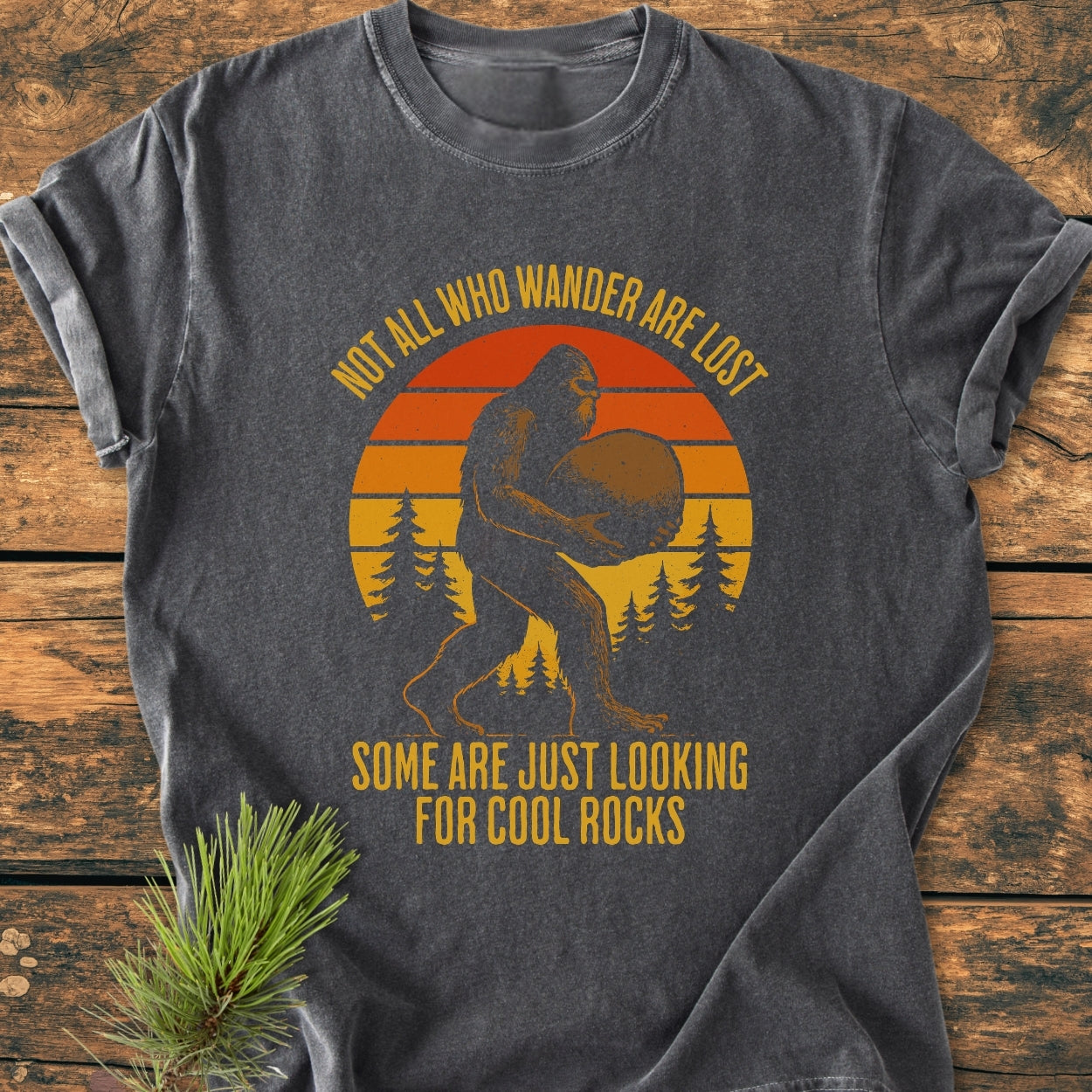 Looking For Cool Rocks - Vintage Wash Tee features Bigfoot under an orange sunset, pine trees, and a witty quote on a dark-gray, soft, relaxed-fit, 100% cotton T-shirt, displayed on wood.