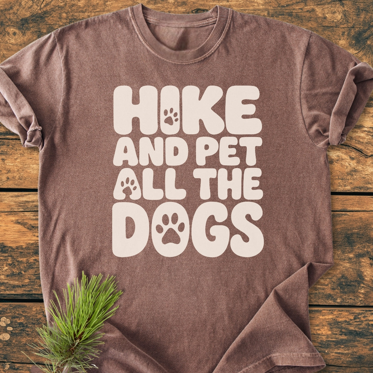Hike And Pet All The Dogs - Vintage Wash Tee, featuring a cream slogan with paw prints on a brown shirt, laid flat on a wooden surface with a small evergreen pine sprig beside it.