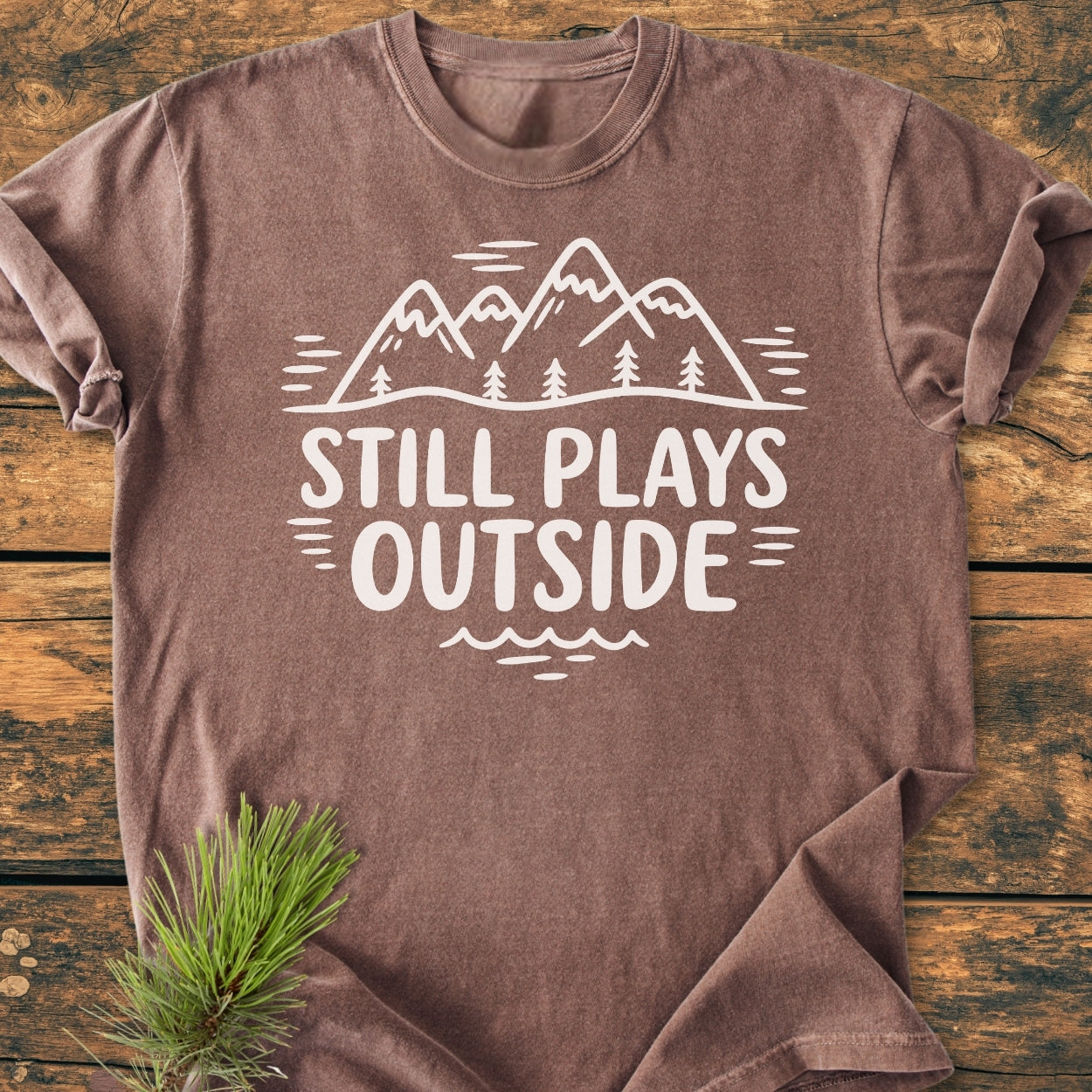 Still Plays Outside - Vintage Wash Tee featuring a stylized mountain range and slogan, laid flat on wood surface with pine sprig accentuating the outdoor theme. Made of soft, durable ring-spun cotton.
