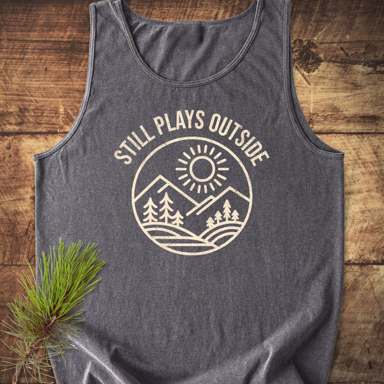 Still Plays Outside Tank Top, charcoal-gray, features a circular outdoor scene print. Laid on rustic wood, it embodies a relaxed fit and sustainable fashion, reinforced by a pine sprig accent.