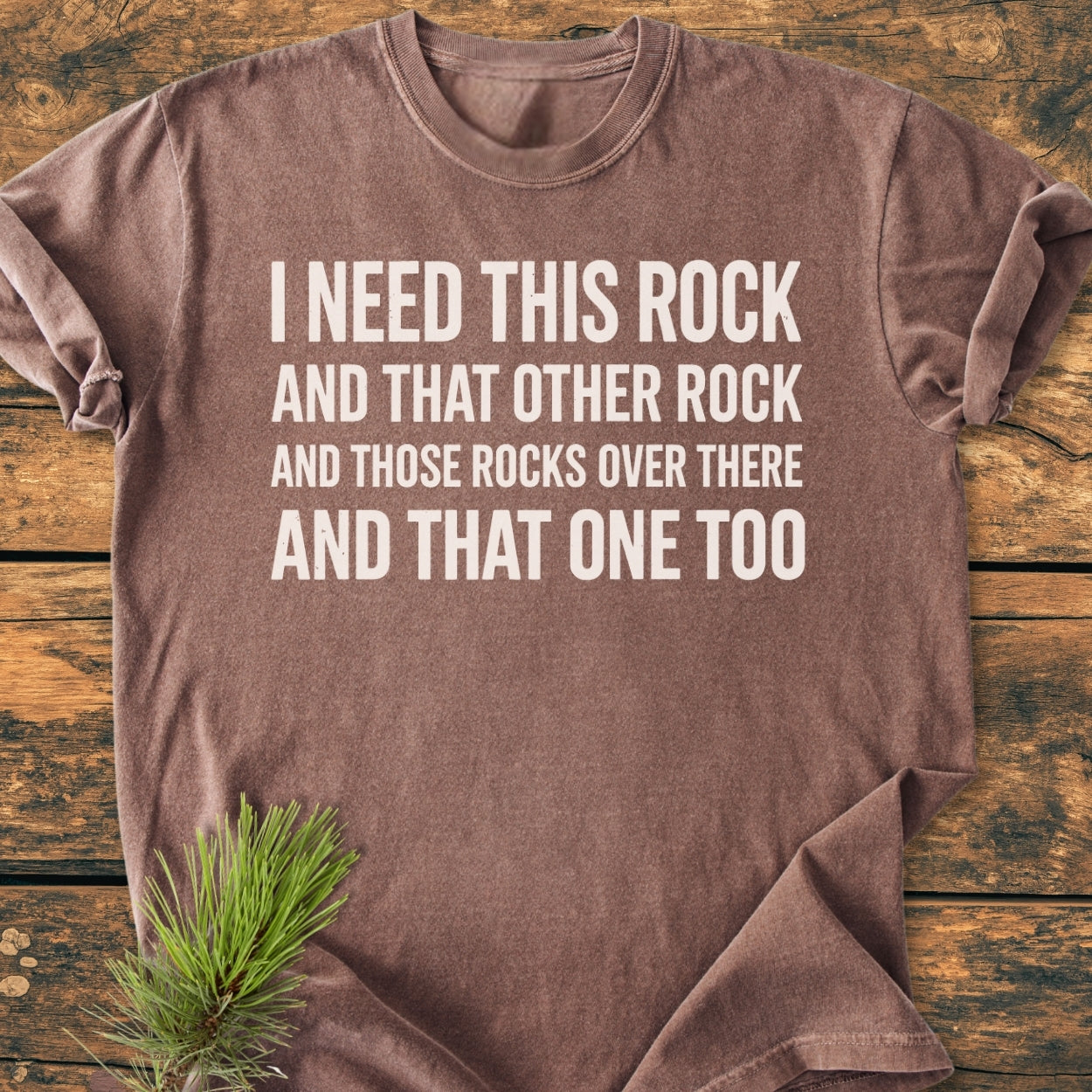 I Need This Rock - Vintage Wash Tee: A brown, short-sleeved T-shirt with bold white text, styled with a pine sprig on a wood surface; made of soft, durable, garment-dyed cotton for comfort.