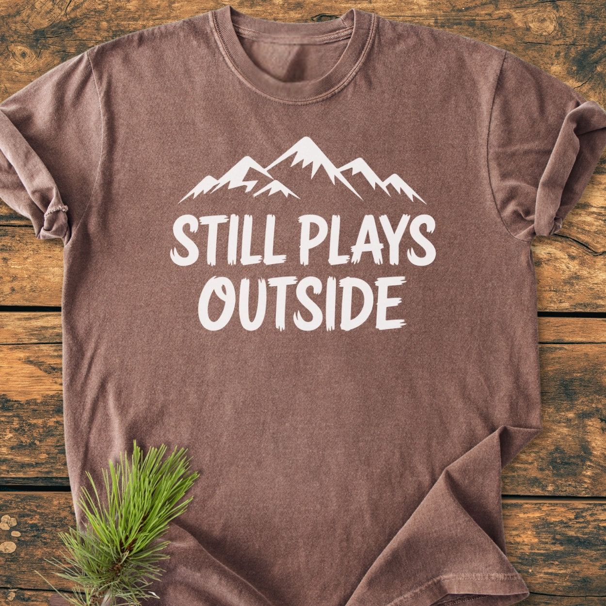 Still Plays Outside - Vintage Wash Tee featuring a mountain silhouette and text, laid flat with a pine sprig accent. Crafted from soft, durable, pre-shrunk 100% ring-spun cotton for everyday comfort.