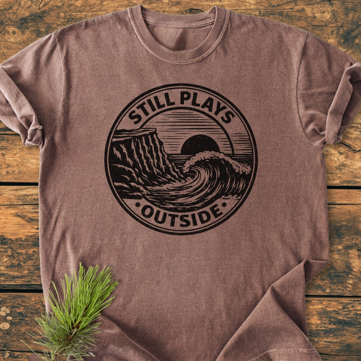 Still Plays Outside - Vintage Wash Tee laid flat on wood, showing a heathered brown color, vintage-style ocean wave graphic, and evergreen sprig beside it, highlighting its relaxed fit and soft, durable fabric.