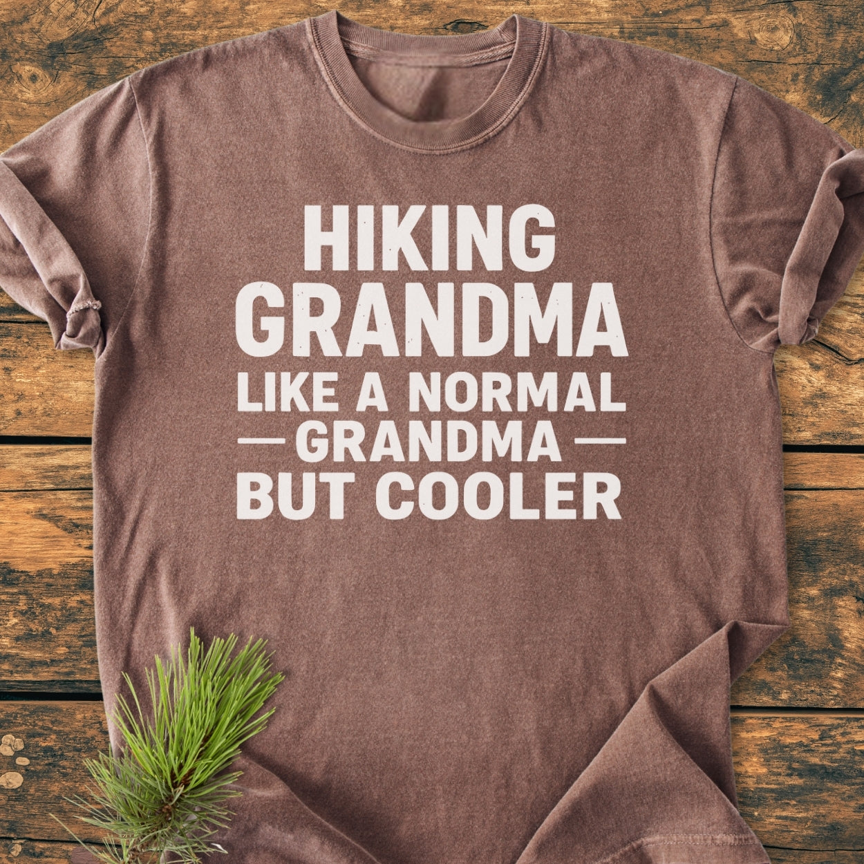 Heather-brown Hiking Grandma - Vintage Wash Tee with bold white slogan, laid on wood, features a pine sprig. Soft, durable, and relaxed fit made of 100% ring-spun cotton.