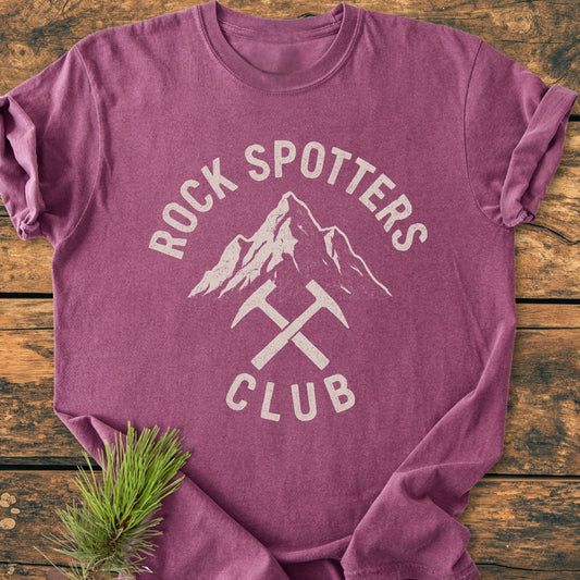 Rock Spotters Club - Vintage Wash Tee in heathered-purple, featuring a white distressed mountain and rock-hammers graphic, laid flat on wood with a pine sprig accentuating its rustic charm.