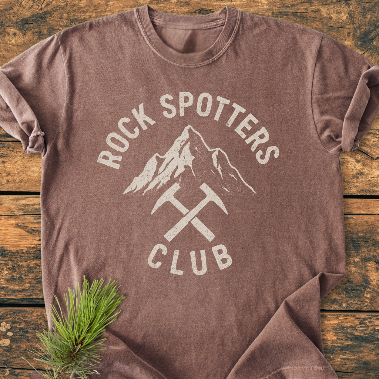 Rock Spotters Club - Vintage Wash Tee features a mountain graphic with crossed rock-hammers on a heather-brown T-shirt, laid flat with a pine sprig, exuding a rustic, outdoorsy vibe.