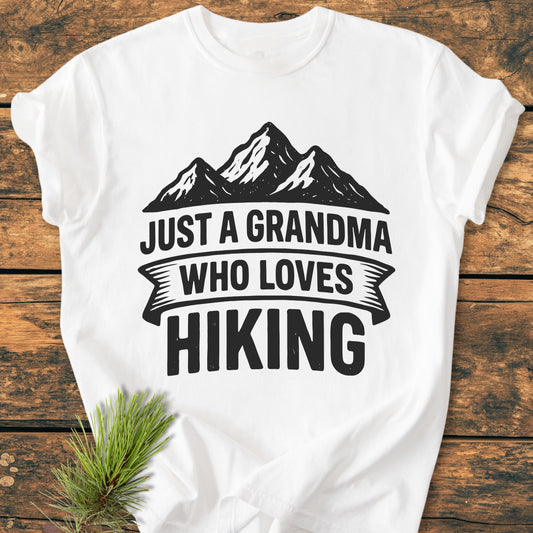 Just A Grandma Who Loves Hiking - Vintage Wash Tee, featuring a mountain silhouette print on a white shirt, laid on wood with a pine sprig, emphasizing comfort and durability with a relaxed fit.