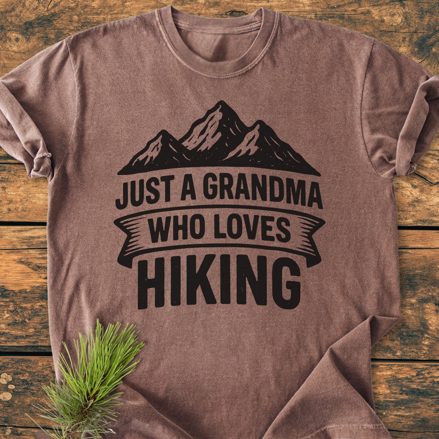 Just A Grandma Who Loves Hiking - Vintage Wash Tee: Heather-brown T-shirt featuring a mountain silhouette and bold lettering, laid flat on a wooden surface with a pine needle sprig accent.