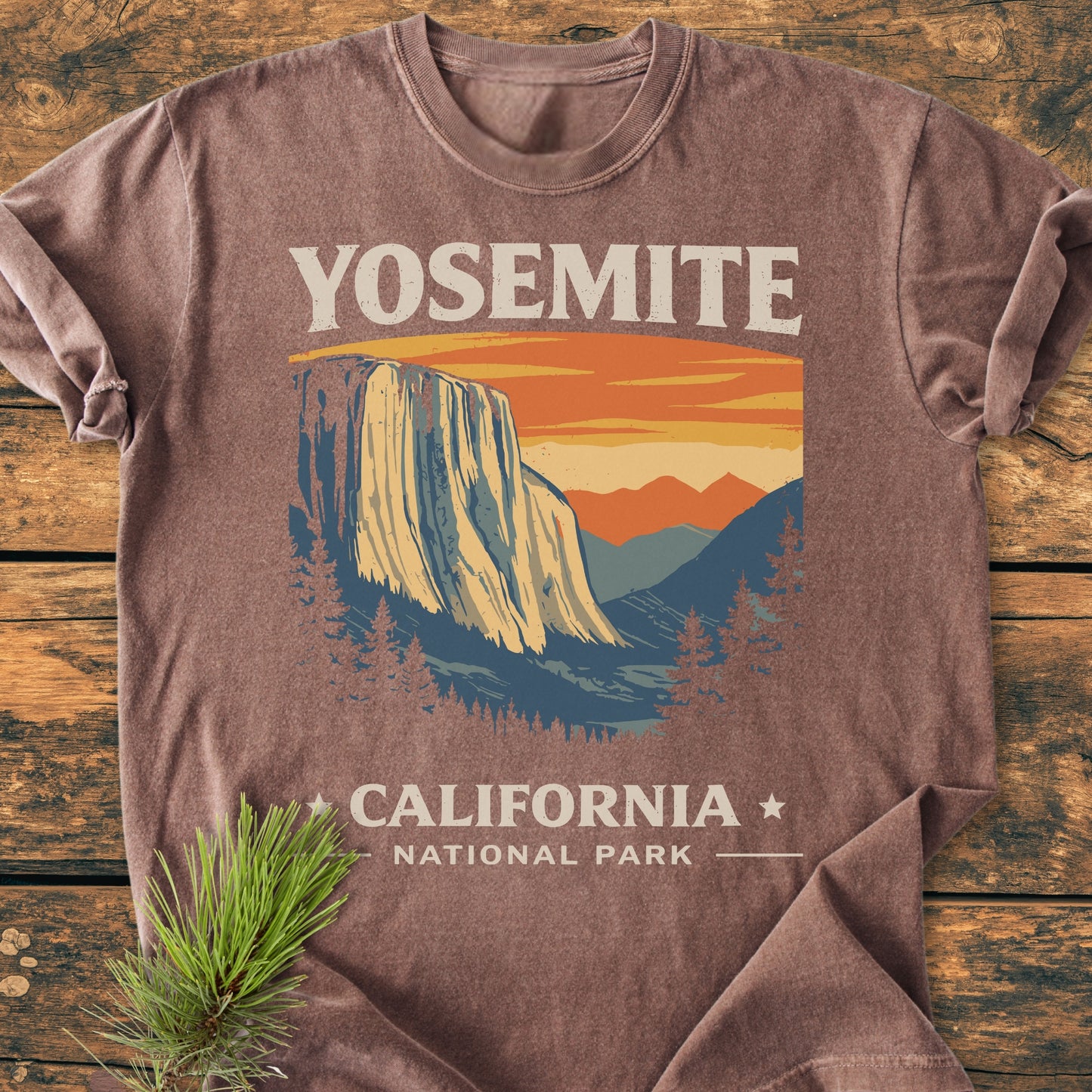 Yosemite National Park - Vintage Wash Tee features a stylized Yosemite design with El Capitan, mountains, and pines, printed on a soft, relaxed-fit, brown vintage-washed T-shirt, laid on a wooden surface.
